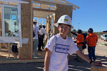 A boy in a hard hat and goggles stands in front of a house under construction