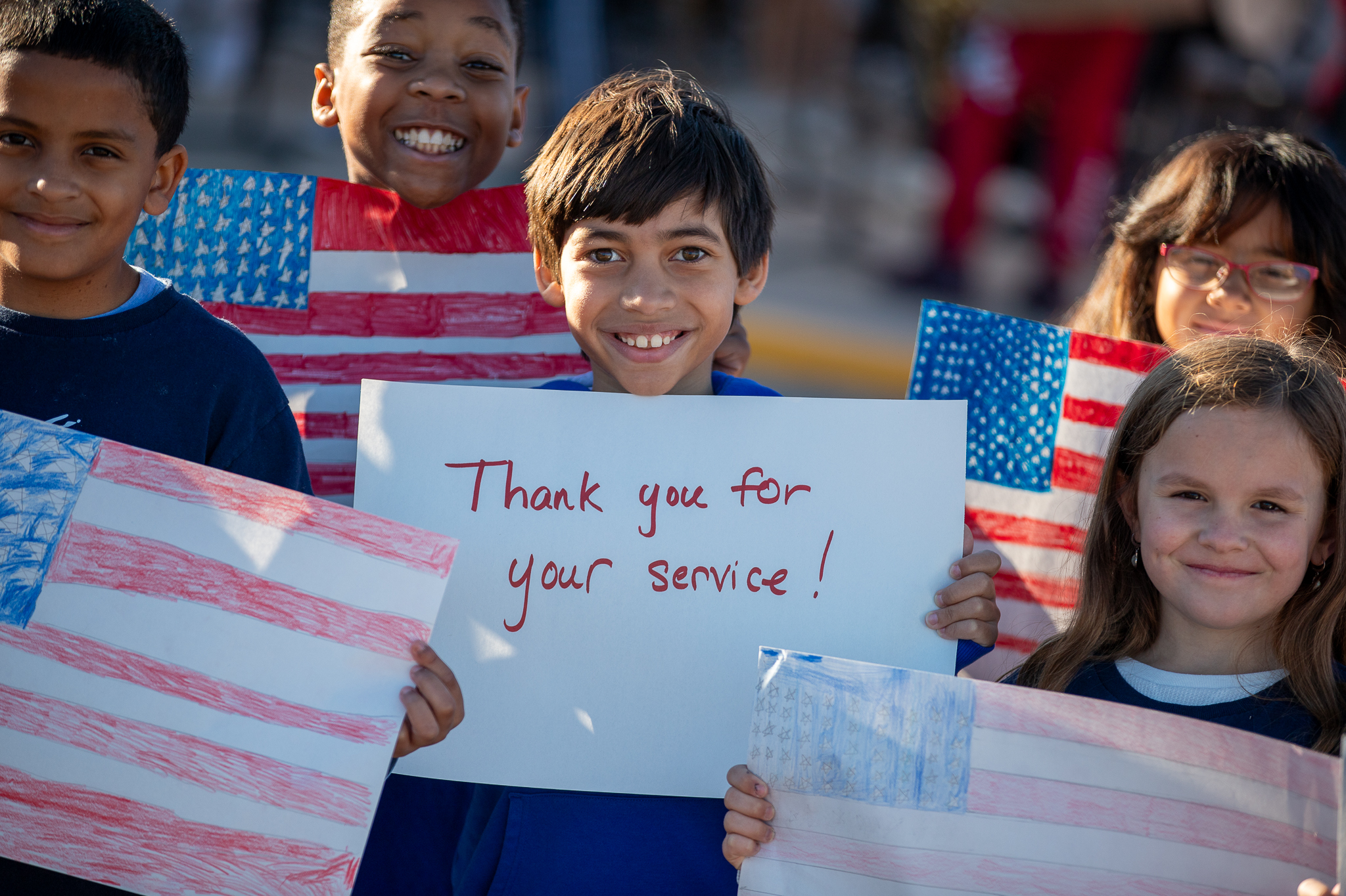 Students hold up hand drawn American flags and a sign reading Thank you for your service!