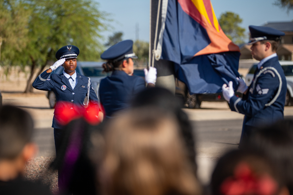 A woman in a blue military uniform salutes the flag as two other people in military uniforms unfold it