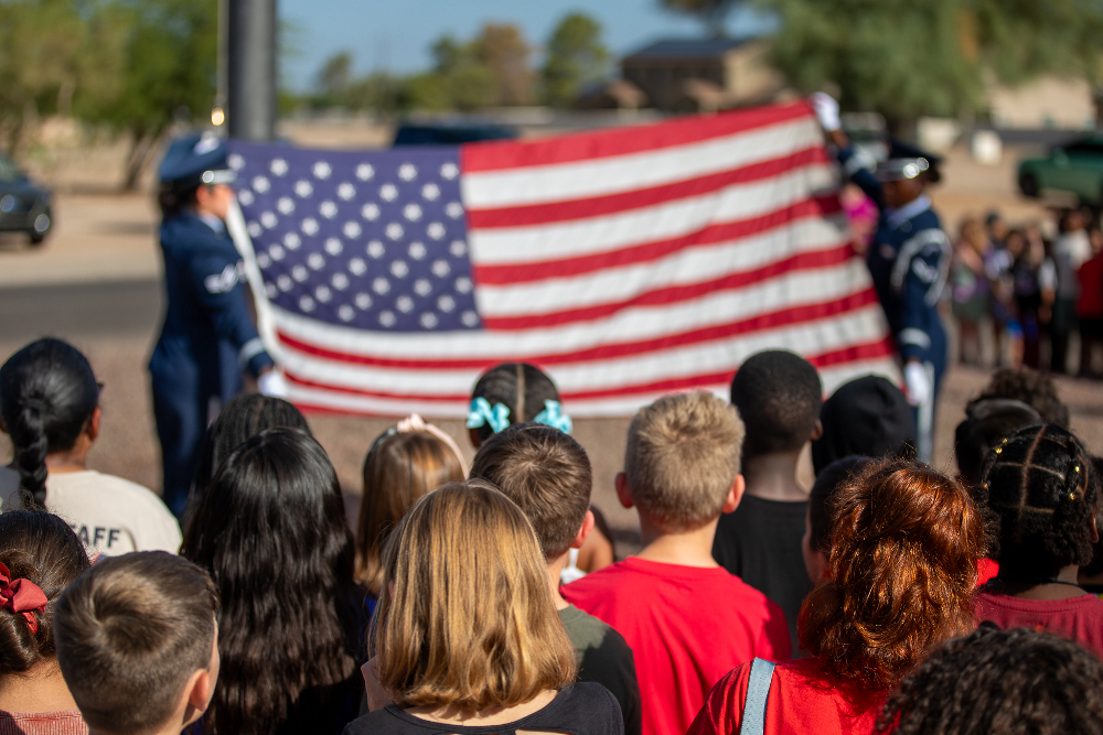 Two women in military uniforms raise the American flag as students watch