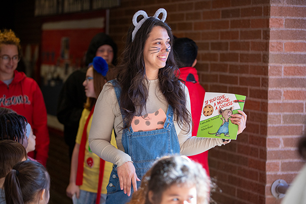 A woman dressed up as a mouse in overalls holds the book If You Give a Mouse a Cookie