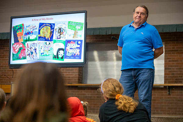 A man in a blue shirt, right, stands in front of a screen with photos of his picture books, as he talks to students