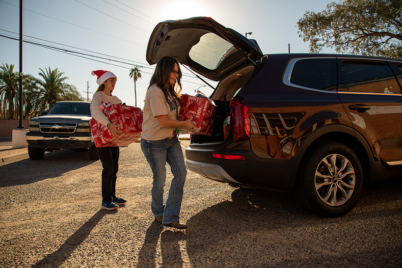 Two women unload wrapped boxes from the trunk of an SUV
