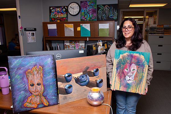 A woman smiles as she holds up a colorful painting of a lion, alongside other artwork